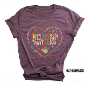 May include: Heather maroon t-shirt with a circle design that says "Inclusion Matters" in colorful letters. The circle is surrounded by words like "diversity", "compassion", "empathy", "kindness", "respect", "ability", "potential", "bravery", "joy", "truth", and "educate".