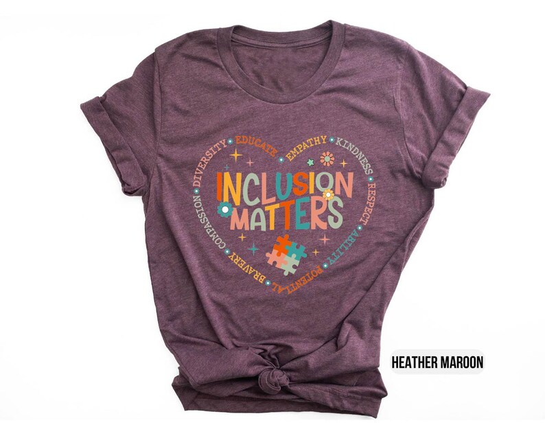 May include: Heather maroon t-shirt with a circle design that says "Inclusion Matters" in colorful letters. The circle is surrounded by words like "diversity", "compassion", "empathy", "kindness", "respect", "ability", "potential", "bravery", "joy", "truth", and "educate".