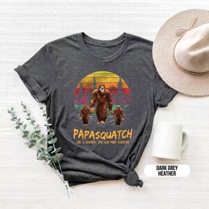 May include: Dark grey heather t-shirt featuring a Bigfoot family graphic. The design includes a large Bigfoot leading two smaller ones, set against a sunset backdrop with the text "PAPASQUATCH". A hat and mug are also in the image.