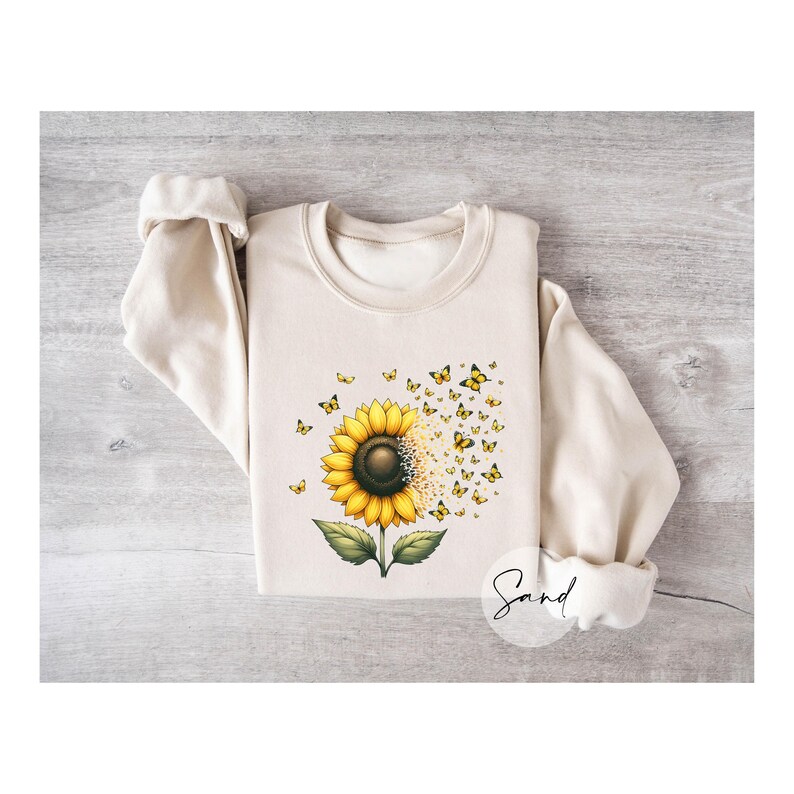 K&ouml;nnte beinhalten: Ein cremefarbener Pullover mit einem Aufdruck einer Sonnenblume mit gelben Bl&uuml;tenbl&auml;ttern und einer braunen Mitte. Schmetterlinge fliegen aus der Sonnenblume, die auf einem gr&uuml;nen Stiel mit Bl&auml;ttern sitzt. Das Wort "Sand" ist in Kursivschrift in der rechten unteren Ecke des Pullovers geschrieben.
