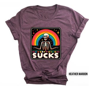 Pu&ograve; includere: Una t-shirt color marrone rossiccio con una grafica di uno scheletro davanti a un arcobaleno con il testo "EVERYTHING SUCKS".