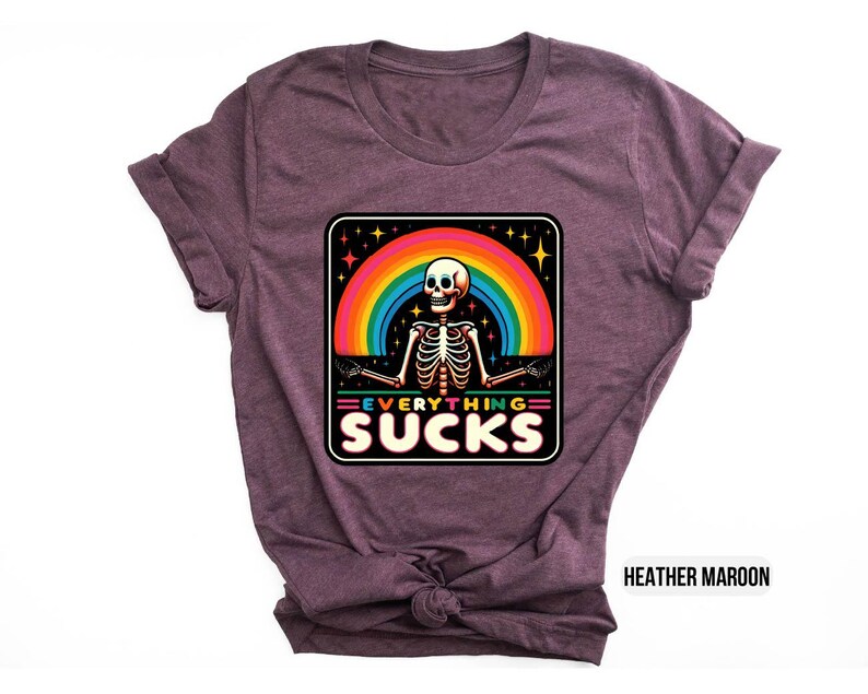 Pu&ograve; includere: Una t-shirt color marrone rossiccio con una grafica di uno scheletro davanti a un arcobaleno con il testo "EVERYTHING SUCKS".