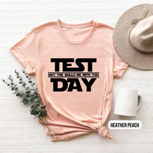 K&ouml;nnte beinhalten: Ein pfirsichfarbenes T-Shirt mit schwarzem Text, der "TEST MAY THE SKILLS BE WITH YOU DAY" lautet.