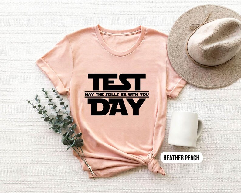 K&ouml;nnte beinhalten: Ein pfirsichfarbenes T-Shirt mit schwarzem Text, der "TEST MAY THE SKILLS BE WITH YOU DAY" lautet.