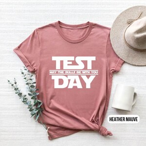 K&ouml;nnte beinhalten: Ein T-Shirt in Heide-Mauve mit dem Text "TEST MAY THE SKILLS BE WITH YOU DAY" in Wei&szlig;.