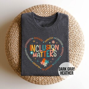 May include: Dark gray heather t-shirt with a colorful graphic of a heart with the words "Inclusion Matters" surrounded by words like "diversity", "empathy", "kindness", and "respect".