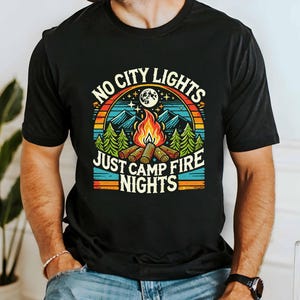 May include: Black t-shirt with a retro-style graphic design featuring a campfire scene and the text "No City Lights Just Camp Fire Nights".