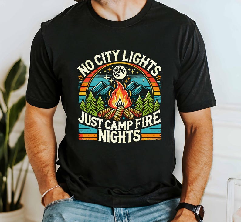 K&ouml;nnte beinhalten: Schwarzes T-Shirt mit einem Retro-Grafikdesign, das eine Feuerstellenszene und den Text "No City Lights Just Camp Fire Nights" zeigt.