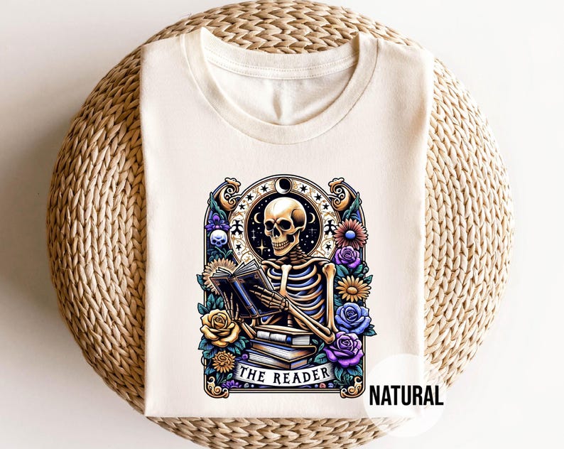 May include: A white t-shirt with a graphic of a skeleton reading a book. The graphic is surrounded by flowers and the text "The Reader" is below the skeleton. The text "NATURAL" is in the bottom right corner of the image.