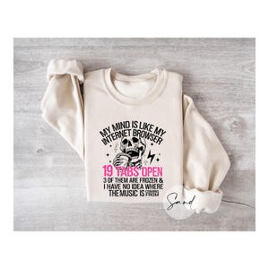 May include: A beige sweatshirt with a black and white graphic of a skull drinking coffee. The text on the sweatshirt reads "My mind is like my internet browser 19 tabs open 3 of them are frozen & I have no idea where the music is from".