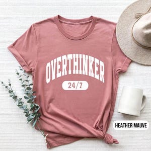 Peut inclure: Un t-shirt mauve clair avec le texte "Overthinker 24/7" en blanc.