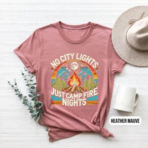 K&ouml;nnte beinhalten: Heather-mauvefarbenes T-Shirt mit einer Grafik im Retro-Stil, auf der "No City Lights Just Camp Fire Nights" steht. Die Grafik zeigt ein Lagerfeuer, B&auml;ume und einen Mond.