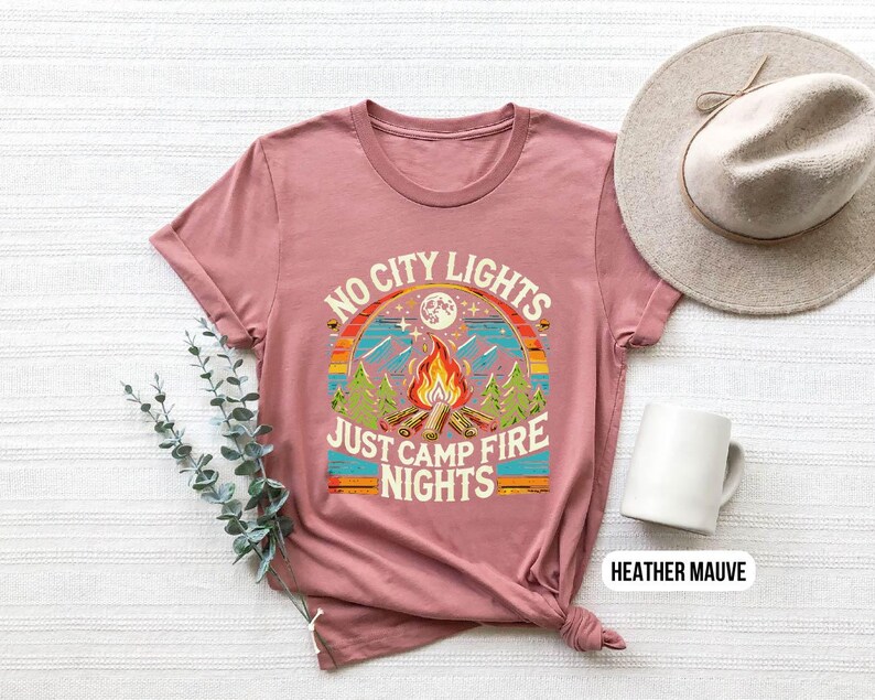 K&ouml;nnte beinhalten: Heather-mauvefarbenes T-Shirt mit einer Grafik im Retro-Stil, auf der "No City Lights Just Camp Fire Nights" steht. Die Grafik zeigt ein Lagerfeuer, B&auml;ume und einen Mond.