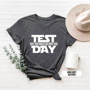 K&ouml;nnte beinhalten: Dunkelgraues T-Shirt mit Melange-Effekt und wei&szlig;em Text, der "TEST MAY THE SKILLS BE WITH YOU DAY" lautet.