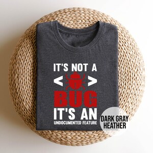 May include: Dark gray heather t-shirt with the text "IT'S NOT A BUG IT'S AN UNDOCUMENTED FEATURE". The word "BUG" is in red with a bug graphic. The shirt is folded on a woven, circular surface.