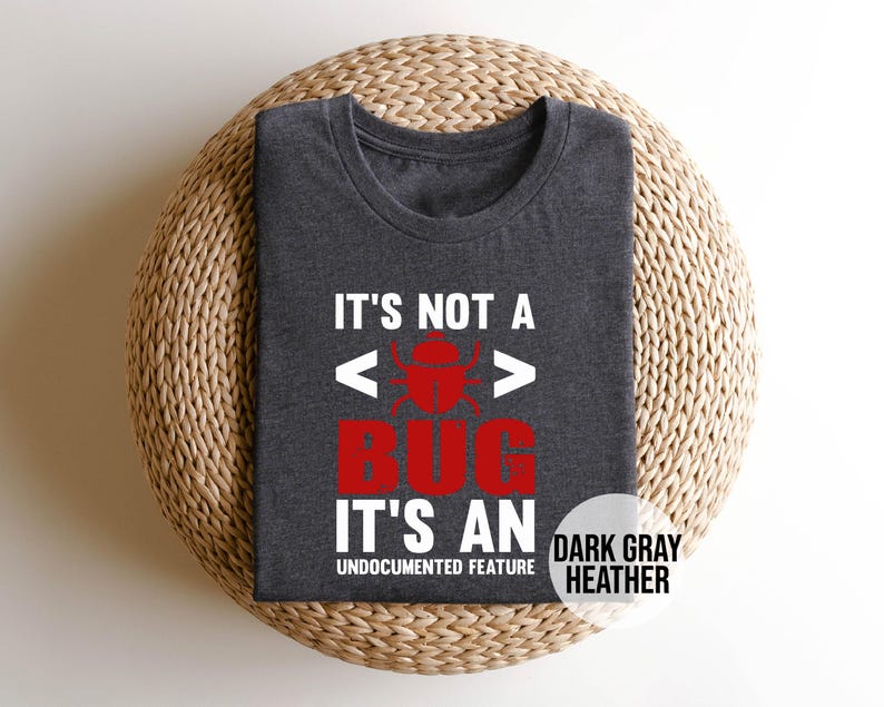 May include: Dark gray heather t-shirt with the text "IT'S NOT A BUG IT'S AN UNDOCUMENTED FEATURE". The word "BUG" is in red with a bug graphic. The shirt is folded on a woven, circular surface.