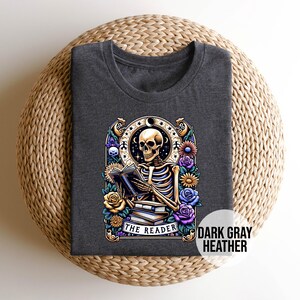 May include: Dark gray heather t-shirt with a graphic of a skeleton reading a book surrounded by flowers. The text "The Reader" is below the skeleton.