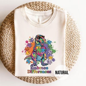 May include: A white t-shirt with a colorful dinosaur holding a puzzle piece heart. The dinosaur is wearing sunglasses and the text "Embrace Differences" is printed below the dinosaur. The word "NATURAL" is printed in the bottom right corner of the shirt.
