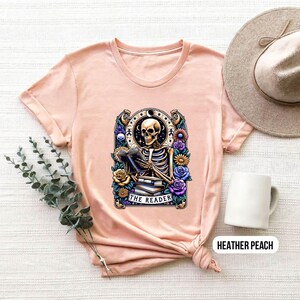 May include: A peach colored t-shirt with a graphic of a skeleton reading a book. The graphic is surrounded by flowers and the text "The Reader".