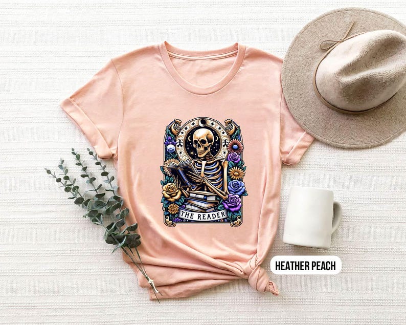 May include: A peach colored t-shirt with a graphic of a skeleton reading a book. The graphic is surrounded by flowers and the text "The Reader".