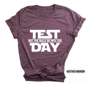 K&ouml;nnte beinhalten: T-Shirt in Heidekraut-Maroon mit wei&szlig;em Text, der "TEST MAY THE SKILLS BE WITH YOU DAY" lautet.
