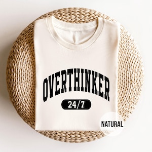 Peut inclure: Un t-shirt blanc avec du texte noir qui dit "Overthinker 24/7" et "Natural" dans une arche noire. Le t-shirt est sur un fond de paille tiss&eacute;e.
