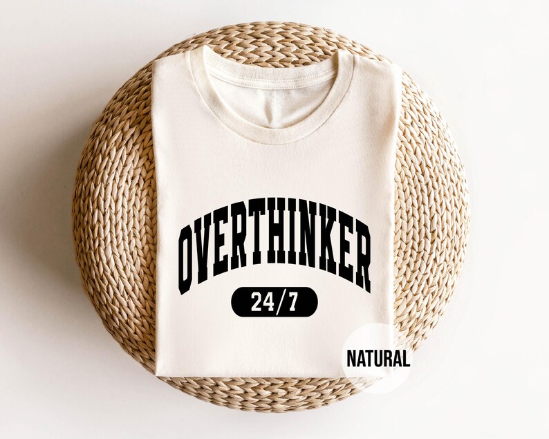 Peut inclure: Un t-shirt blanc avec du texte noir qui dit "Overthinker 24/7" et "Natural" dans une arche noire. Le t-shirt est sur un fond de paille tiss&eacute;e.