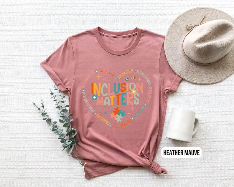 May include: Heather mauve t-shirt with a heart-shaped graphic that says "Inclusion Matters" surrounded by words like "empathy", "kindness", "respect", and "educate".