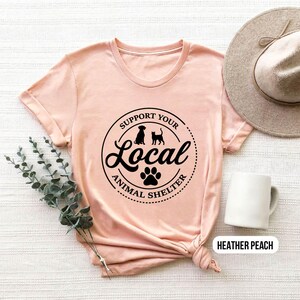 May include: A heather peach t-shirt with a black graphic that says "Support Your Local Animal Shelter" with a paw print and silhouettes of a dog and cat.