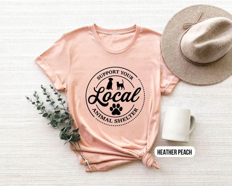 May include: A heather peach t-shirt with a black graphic that says "Support Your Local Animal Shelter" with a paw print and silhouettes of a dog and cat.