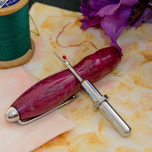 May include: A close-up of sewing tools and supplies. A wooden spool of green thread sits next to a seam ripper with a burgundy handle and silver accents. The seam ripper rests on a piece of fabric with a floral pattern.