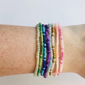 May include: A collection of beaded bracelets in various colors, including green, blue, purple, white, and pink. The bracelets are made of small, round beads strung together. The bracelets are stacked on top of each other.
