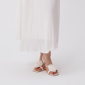 Op de afbeelding: Witte sandalen met hoge hakken, een vierkante neus en een grote stoffen roos op de teenband. De sandalen hebben een dunne hak en een witte enkelband. Gedragen met een witte geplooide rok.