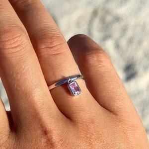Puede incluir: Un anillo de plata con un pequeño colgante rectangular de piedra rosa. El anillo es sencillo y delicado, con una banda fina. La piedra preciosa es de color rosa claro y cuelga de la banda del anillo.