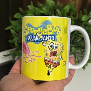 May include: Yellow ceramic mug featuring the text "SpongeBob SquarePants" and an image of SpongeBob and a jellyfish. The mug has a white handle and is held in a hand.