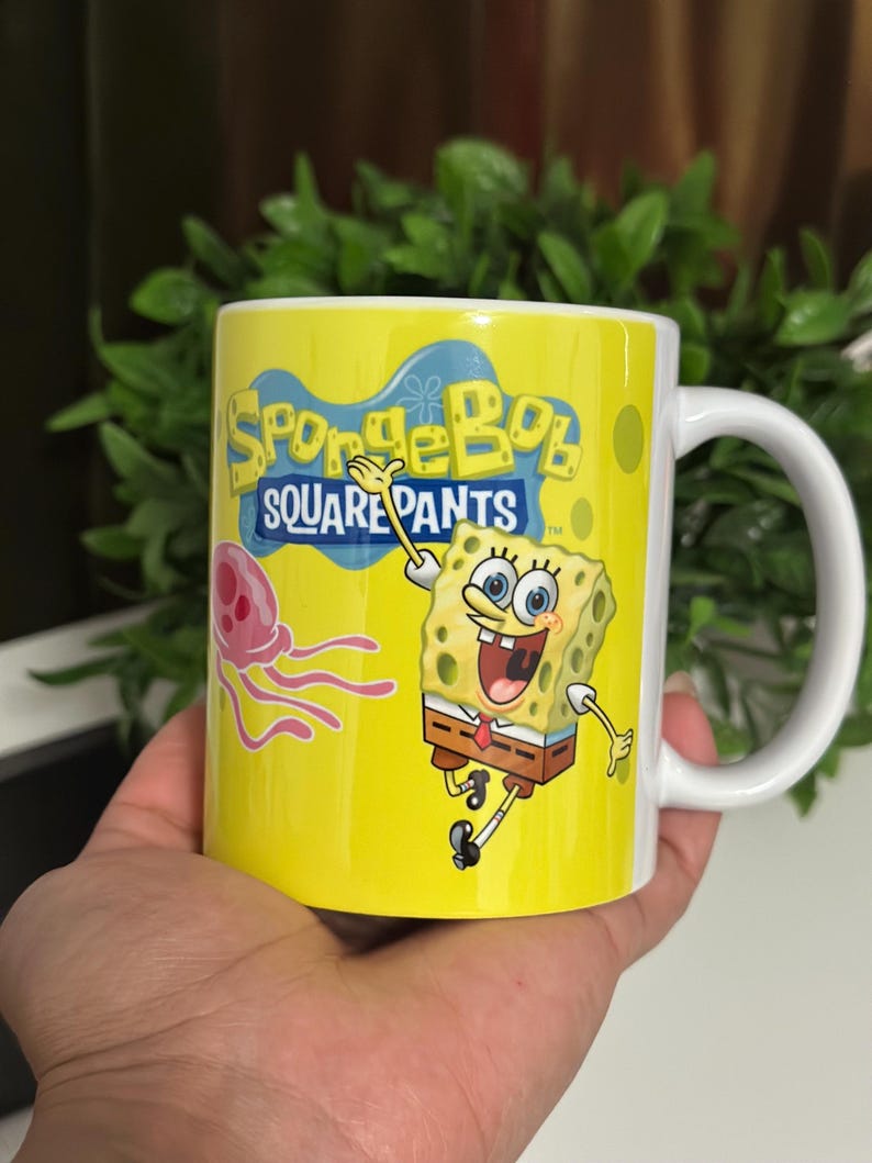 May include: Yellow ceramic mug featuring the text "SpongeBob SquarePants" and an image of SpongeBob and a jellyfish. The mug has a white handle and is held in a hand.
