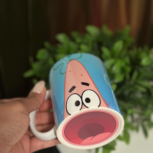 May include: A ceramic mug featuring a cartoon character with a surprised expression. The mug has a light blue background with the character's face and open mouth prominently displayed. The handle is white.