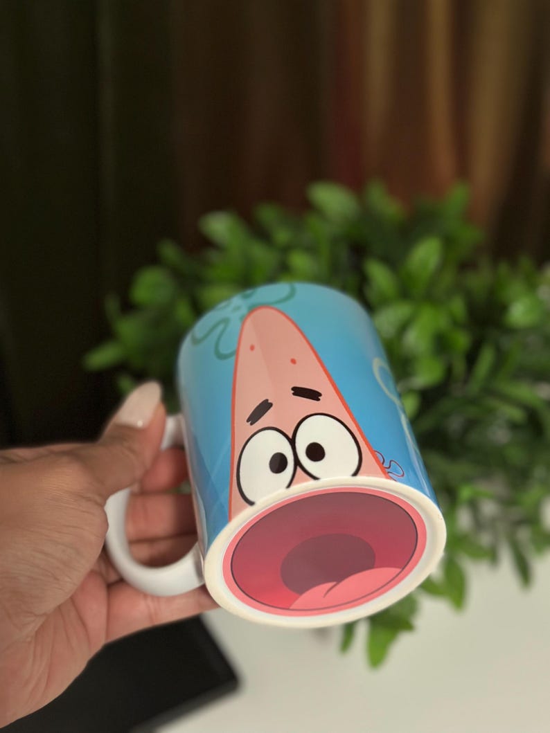 May include: A ceramic mug featuring a cartoon character with a surprised expression. The mug has a light blue background with the character's face and open mouth prominently displayed. The handle is white.
