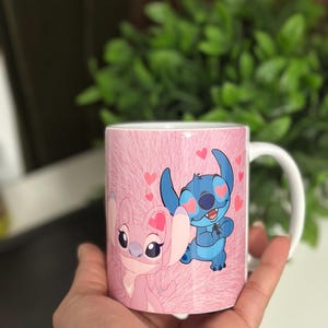 May include: A white ceramic mug featuring a pink background with a pattern. The mug is decorated with cartoon characters, a pink one and a blue one, with hearts. The mug is held by a hand.