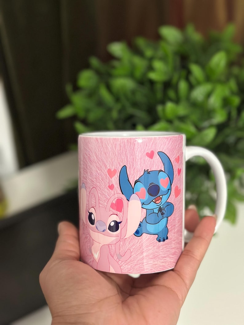 May include: A white ceramic mug featuring a pink background with a pattern. The mug is decorated with cartoon characters, a pink one and a blue one, with hearts. The mug is held by a hand.