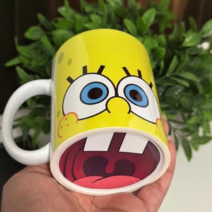 May include: Yellow ceramic mug featuring a cartoon character's face with large blue eyes, a wide open mouth, and a white handle. The mug has a yellow background with small brown spots.