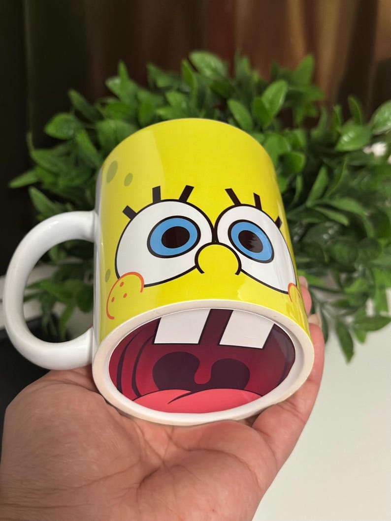 May include: Yellow ceramic mug featuring a cartoon character's face with large blue eyes, a wide open mouth, and a white handle. The mug has a yellow background with small brown spots.