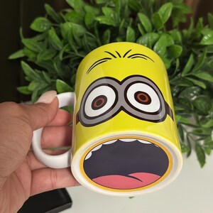 May include: Yellow ceramic mug featuring a Minion character design. The mug has a white handle and a wide open mouth design on the bottom. The Minion's eyes and eyebrows are also visible on the mug.