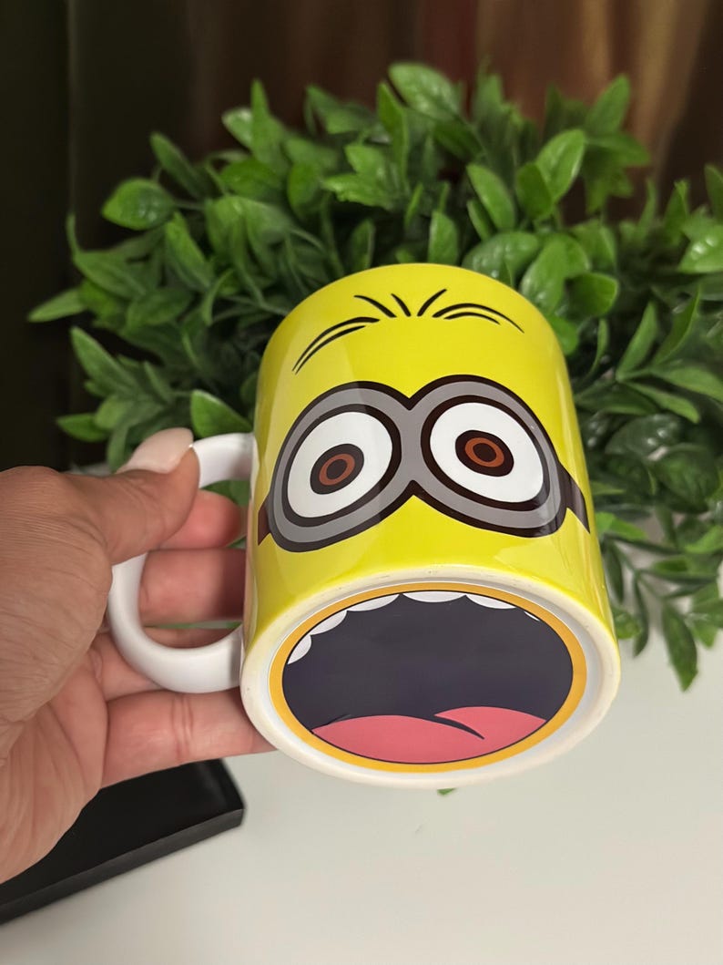 May include: Yellow ceramic mug featuring a Minion character design. The mug has a white handle and a wide open mouth design on the bottom. The Minion's eyes and eyebrows are also visible on the mug.