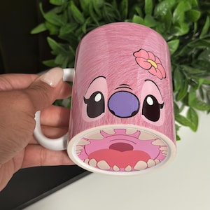 May include: Pink ceramic mug featuring a cartoon character design. The mug has a white handle and a pink exterior with a character's face and a flower. The character's mouth is visible at the bottom of the mug.