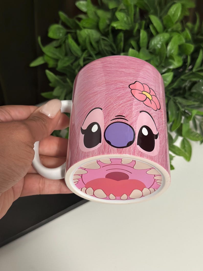 May include: Pink ceramic mug featuring a cartoon character design. The mug has a white handle and a pink exterior with a character's face and a flower. The character's mouth is visible at the bottom of the mug.