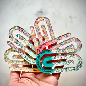 May include: A set of five colorful floral patterned bookmark clips with inspirational quotes. The quotes are: "Trust in the Lord with all your heart", "I am with you", "For I know the plans", "When all", and "Proverbs 3:5".