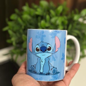 May include: A light blue ceramic mug featuring a cartoon character, Stitch, with large eyes and ears. The mug has a white handle and a blue floral pattern background. The mug is held in a hand.
