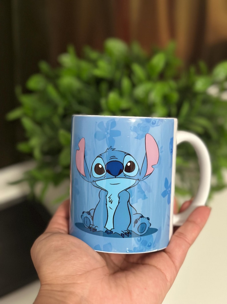May include: A light blue ceramic mug featuring a cartoon character, Stitch, with large eyes and ears. The mug has a white handle and a blue floral pattern background. The mug is held in a hand.