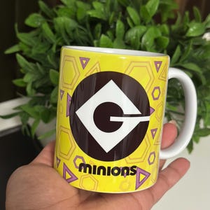 May include: A yellow ceramic mug featuring the Minions logo and the word "minions" in black. The mug has a white handle and is decorated with purple triangles and yellow hexagons. The mug is held in a hand.